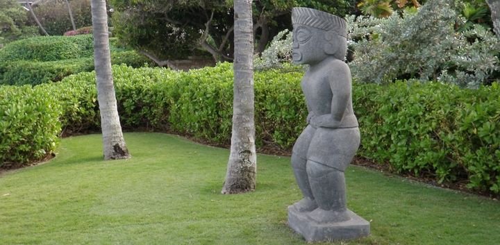 Carved stone figure standing on a lawn between palms, Black Point Residence, Oahu